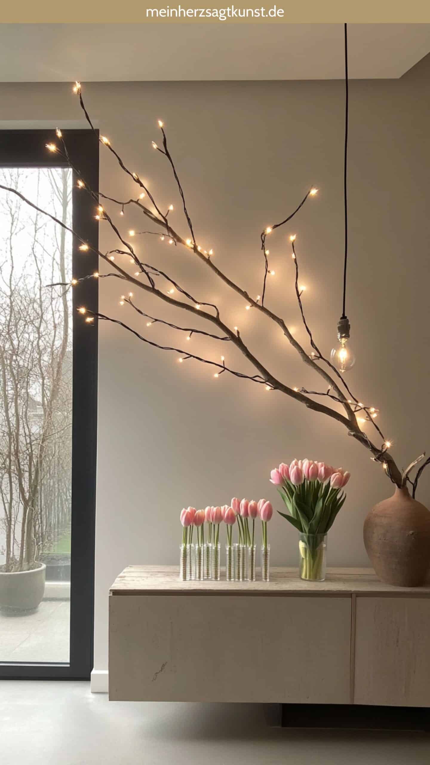 Beleuchteter Ast über Sideboard, darunter Vasengruppe mit Tulpenarrangements